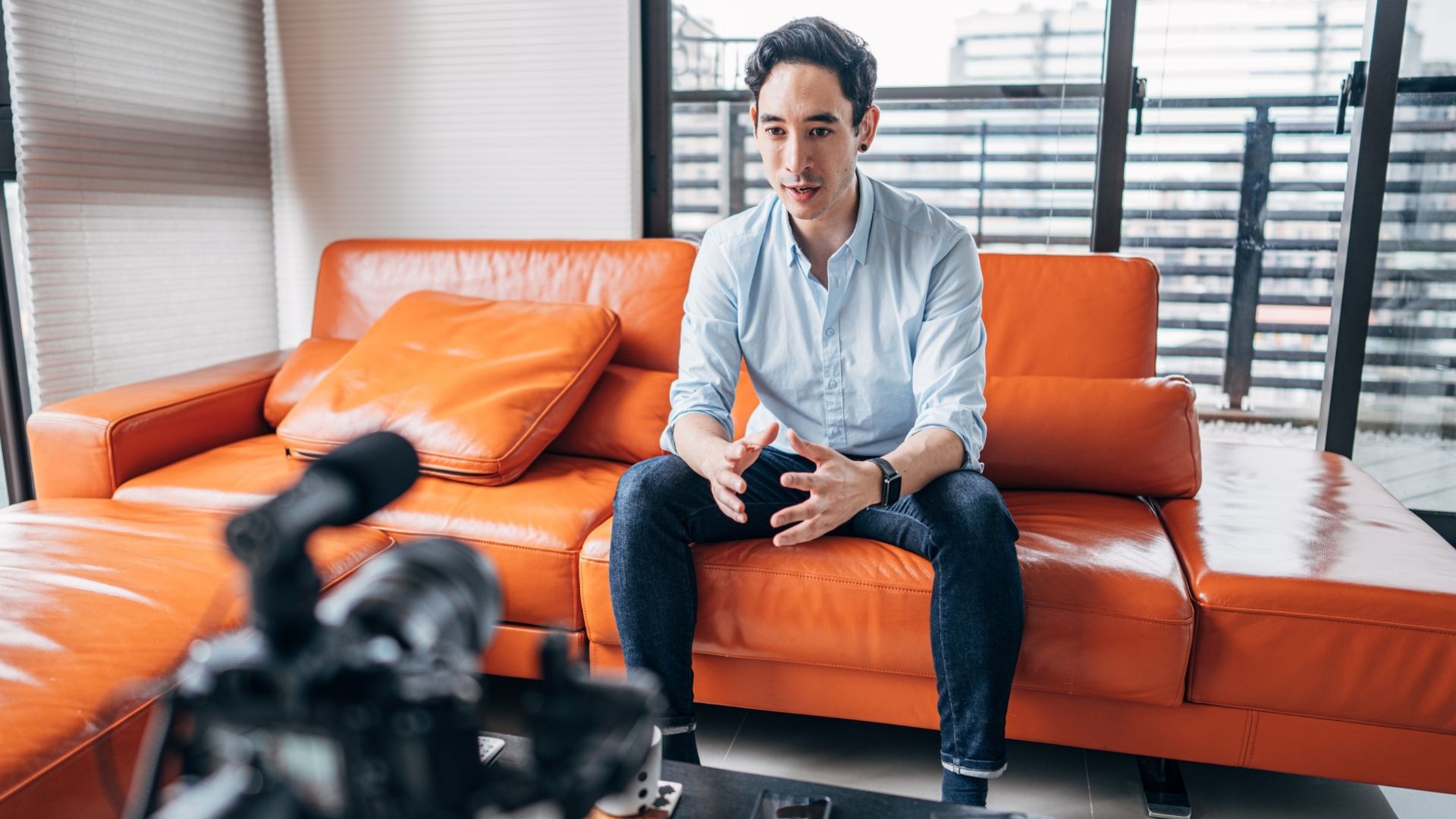 A dark haired man sits on an orange leather sofa making a vlog